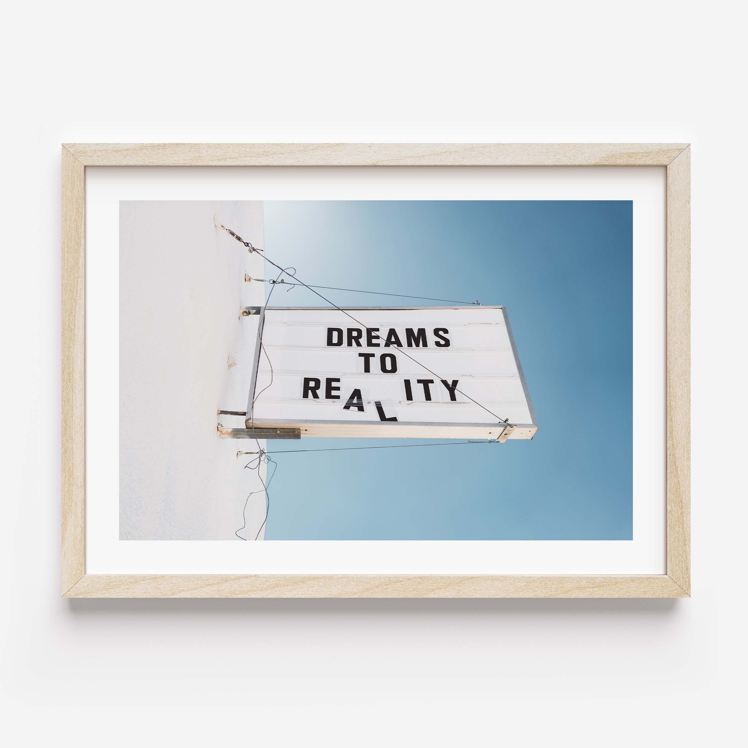 Minimal photo print of a marquee sign on a white wall reading “DREAMS TO REALITY” against a clear blue sky.