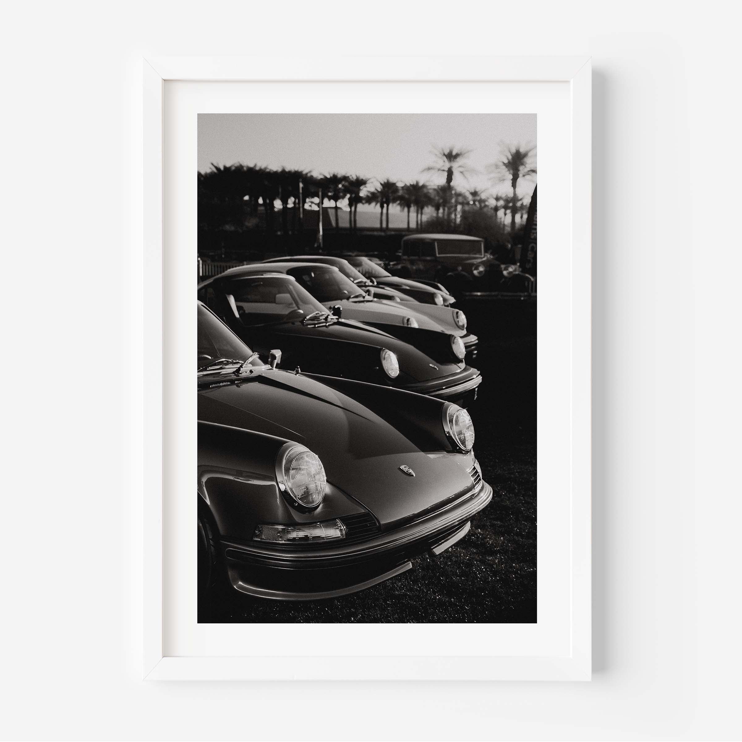 Black-and-white photo print of a row of classic Porsche 911 sports cars parked with palm trees in the background.